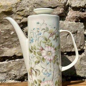 Peut inclure: Une cafetière en céramique blanc cassé avec un motif floral. La cafetière a un long bec verseur, une anse et un couvercle. Le motif floral comprend des roses roses, des campanules bleues et des feuilles vertes. Pot vintage.
