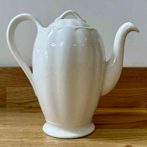 May include: A white ceramic teapot with a curved spout and handle. The teapot has a lid with a small handle and a ribbed design on the body. The teapot is sitting on a wooden surface.