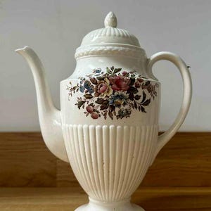 Op de afbeelding: Crèmekleurige keramische koffiepot met een bloemenmotief. De pot heeft een deksel, tuit en handvat. De body heeft verticale geribbelde details. Het bloemmotief bevat roze, blauwe en bruine bloemen.