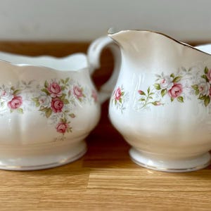 Può includere: Due zuccheriere e lattine in porcellana bianca con un motivo floreale a rose. Le ciotole hanno bordi dorati. Lo zuccheriera è rotonda e la lattiera ha un beccuccio e un manico.