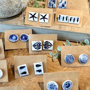 Könnte beinhalten: Sortiment an blauen und weißen Keramikohrringen mit verschiedenen Designs, darunter Seesterne, Fische, Muscheln und Blätter. Die Ohrringe werden auf braunem Karton präsentiert.