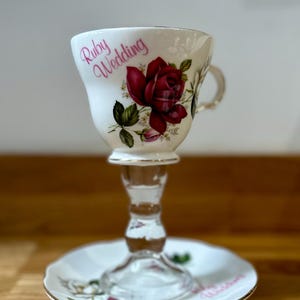 May include: A vintage teacup and saucer set, featuring a large red rose design and the words "Ruby Wedding". The teacup is mounted on a clear glass stem. The saucer and cup have a gold trim.