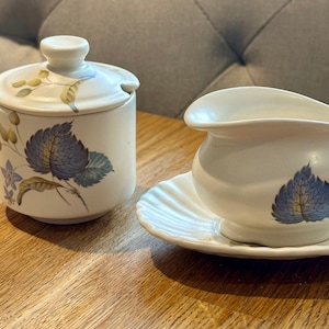 Può includere: Una zuccheriera in ceramica bianca con coperchio e una cremiera e un piattino abbinati. Il set presenta un design floreale blu e verde.