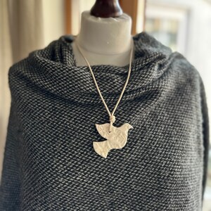 May include: A white ceramic dove pendant on a white cord necklace. The necklace is worn by a mannequin wearing a gray sweater.