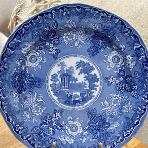 Puede incluir: Un plato de porcelana azul y blanco con un diseño floral y una escena central que representa un edificio con árboles y figuras. El plato tiene un borde festoneado.