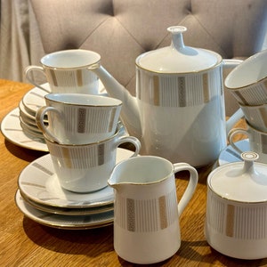 Może przedstawiać: Biały porcelanowy zestaw do herbaty ze złotymi akcentami. Zestaw zawiera czajnik, cukiernicę, dzbanek do śmietanki i sześć filiżanek i spodków. Filiżanki i spodki mają delikatny kwiatowy wzór.