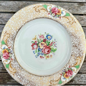 Könnte beinhalten: Ein weißer Keramik-Dinnerteller mit einem goldenen und grünen Blumenmuster. Der Teller hat eine weiße Mitte mit einem Strauß aus rosa, gelben und blauen Blumen. Der Rand des Tellers ist mit einem goldenen und grünen Blumenmuster verziert.