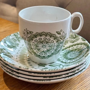 May include: A white teacup with a handle and a matching saucer set. The cup and saucers feature a green floral pattern. The teacup is sitting on top of three additional saucers, all stacked on a wooden surface.