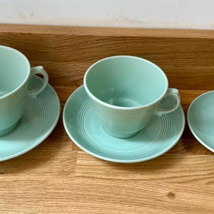 Puede incluir: Tres tazas de té y platillos de color aguamarina están dispuestas sobre una superficie de madera. Las tazas tienen asas y se asientan sobre platillos a juego con diseños de círculos concéntricos. El juego incluye tamaños de taza variables.