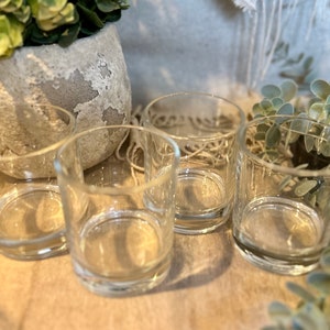 May include: Four clear glass votive candle holders. The cylindrical holders are empty and sit on a neutral-toned surface. The background includes a decorative pot with greenery and additional foliage.