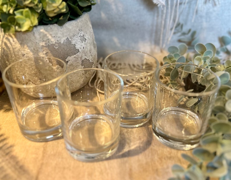 May include: Four clear glass votive candle holders. The cylindrical holders are empty and sit on a neutral-toned surface. The background includes a decorative pot with greenery and additional foliage.
