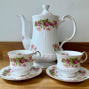 Puede incluir: Un juego de té de porcelana blanca con una tetera, dos tazas y platillos. El juego está decorado con un motivo floral de rosas rosas y flores amarillas. Los bordes y las asas tienen detalles dorados. La tetera tiene un pico largo.