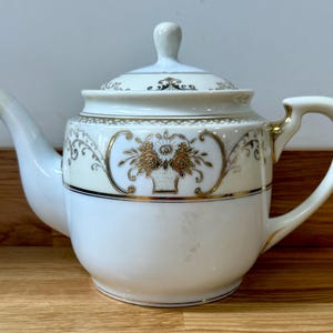 Puede incluir: Tetera de porcelana blanca con tapa, con diseños florales y de volutas doradas. La tetera tiene un pico y un asa curvados, y una banda dorada alrededor del centro. Un pequeño pomo se encuentra en la parte superior de la tapa.