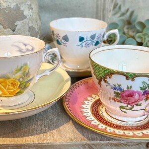 May include: Three antique teacups and saucers. One cup features a yellow rose design, another has a floral pattern, and the third has a green and pink floral design. The saucers are in matching colors.