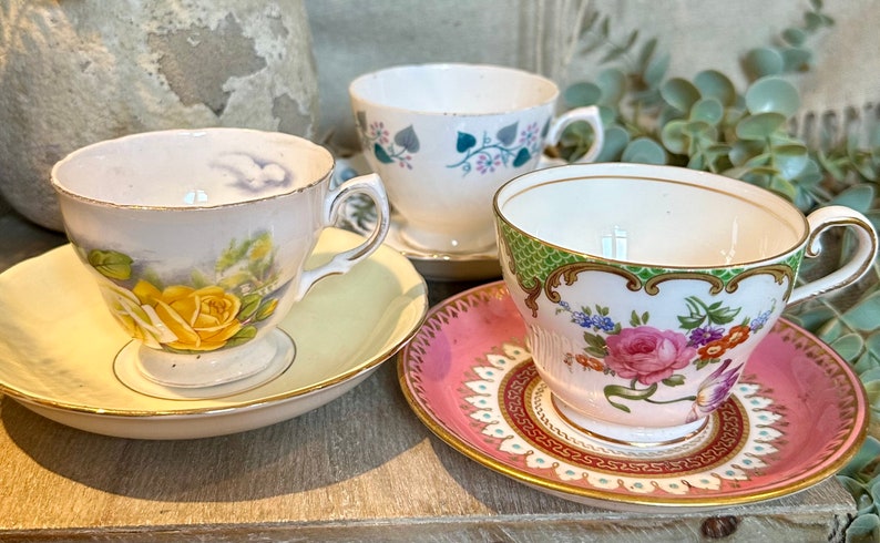 May include: Three antique teacups and saucers. One cup features a yellow rose design, another has a floral pattern, and the third has a green and pink floral design. The saucers are in matching colors.