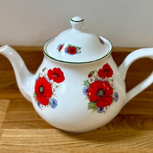 Può includere: Teiera in ceramica bianca con coperchio, con un vivace motivo floreale di papaveri rossi e fiordalisi blu. La teiera ha un beccuccio curvo e un grande manico. Il bordo e il manico sono rifiniti con un bordo verde.