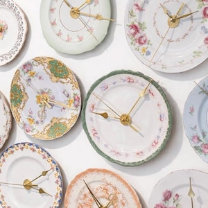 Könnte beinhalten: Eine Sammlung dekorativer Wanduhren aus Vintage-Tellern. Jede Uhr hat ein einzigartiges Blumenmuster und goldfarbene Zeiger. Die Teller variieren in der Farbe, darunter Weiß, Blau und Grün, mit aufwendigen Designs.