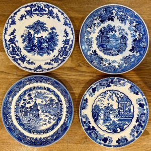 Op de afbeelding: Vier blauwe en witte decoratieve borden met gedetailleerde landschapsscènes. Elk bord heeft een ander ontwerp, waaronder figuren, gebouwen en bloemenranden. De borden zijn gerangschikt op een houten oppervlak.