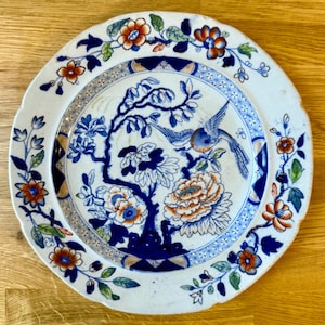 以下が含まれることがあります： 青とオレンジの花と鳥のデザインが施された白いセラミックプレート。プレートの中央には、枝にとまる鳥のイメージがあり、花と葉で囲まれています。縁にはさらに花が飾られています。