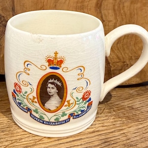 Op de afbeelding: Een crèmekleurige keramische mok met een craquelé glazuurafwerking, voorzien van een centraal ovaal portret van koningin Elizabeth II. Boven het portret staat een kroon, geflankeerd door bloemmotieven in rood, oranje en groen. De tekst luidt Coronation Queen Elizabeth II 2nd June 1953.