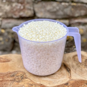 May include: A clear plastic measuring cup filled with off-white, pellet-shaped wax. The cup has a handle and a spout for easy pouring. The wax pellets are tightly packed, suggesting a bulk quantity of candle-making supplies.