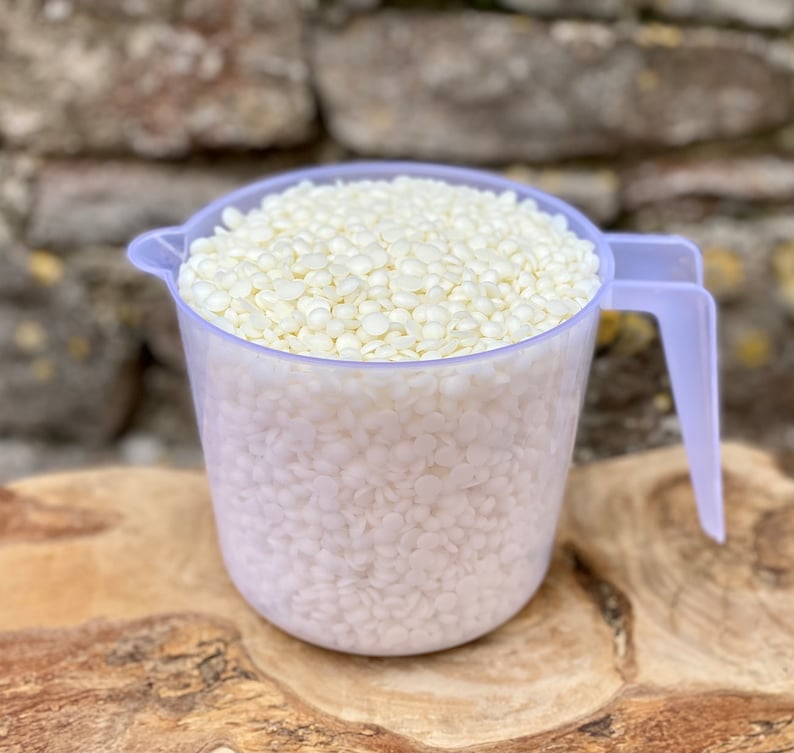 May include: A clear plastic measuring cup filled with off-white, pellet-shaped wax. The cup has a handle and a spout for easy pouring. The wax pellets are tightly packed, suggesting a bulk quantity of candle-making supplies.