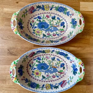 May include: Two oval ceramic serving dishes with integrated handles, featuring a cream-coloured base decorated with a colourful floral pattern of blue, yellow, and pink flowers, green leaves, and a small bird motif, finished with a blue border.