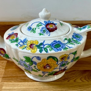 Könnte beinhalten: Cremefarbene Keramik-Teekanne mit Deckel, Ausguss und Henkel. Die Teekanne ist mit bunten Blumenmustern in Rot, Gelb und Blau verziert. Die Teekanne steht auf einer Holzoberfläche. Ca. 20 cm lang.