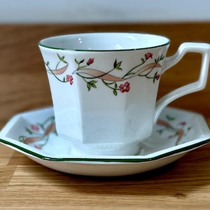 Puede incluir: Juego de taza y platillo blancos con diseño floral. La taza tiene forma geométrica con asa cuadrada y ribete verde. El platillo es cuadrado con ribete verde y estampado floral a juego. El diseño floral incluye flores rosas y una cinta rosa.