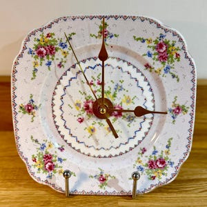 May include: Square decorative clock made from a floral patterned plate. The clock face features a cross-stitch design with pink, blue, and yellow flowers. The clock hands are gold-colored, and the clock is mounted on a metal stand.