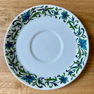 May include: White saucer with a floral design in teal and green. The pattern features stylized flowers and leaves arranged around the rim. The saucer has a central recessed area for a teacup.
