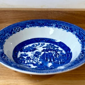 May include: A round ceramic bowl featuring a classic blue and white willow pattern. The design includes intricate illustrations of pagodas, trees, and birds within a central circular medallion, surrounded by a wide, decorative geometric border.