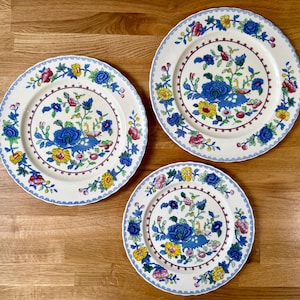 Op de afbeelding: Drie ronde keramische borden met een crèmekleurige basis en een kleurrijk bloemmotief. Het ontwerp bevat blauwe, gele en roze bloemen, groene bladeren en kleine vogelillustraties. De randen hebben een blauw geschulpt patroon.