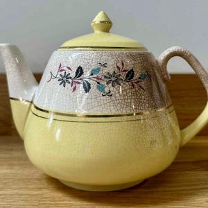 Puede incluir: Una tetera amarilla vintage con un diseño floral. La tetera tiene un pico y asa blancos, y una tapa con un adorno dorado. El diseño floral incluye detalles en negro, rojo y azul.
