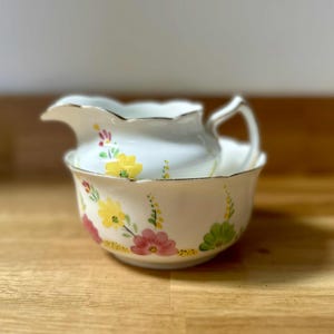 Può includere: Un set di brocca e ciotola in porcellana bianca. Il set è decorato con motivi floreali gialli, rosa e verdi. I bordi di entrambi i pezzi sono rifiniti in oro. La brocca è appoggiata all'interno della ciotola.