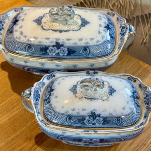 May include: Two rectangular ceramic serving dishes with lids, featuring a white base with intricate blue floral patterns, basket-weave borders, and gold-trimmed edges. Each dish has two side handles and a decorative, ornate handle on the lid.