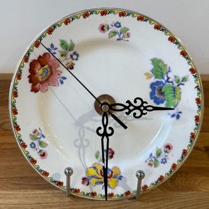 Puede incluir: Un reloj decorativo hecho con un plato. El plato blanco presenta patrones florales coloridos en rojo, azul y amarillo. Las manecillas negras y el centro de latón completan el diseño. El reloj se muestra en un soporte de metal plateado.