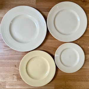 Può includere: Quattro piatti in ceramica color crema di varie dimensioni sono disposti su una superficie di legno. I piatti hanno un design semplice con anelli concentrici attorno al bordo. I piatti sono di colore neutro, adatti a vari contesti di ristorazione.