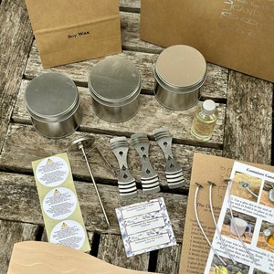 May include: A candle-making kit with various supplies, including soy wax, metal tins, a thermometer, wicks, and a wooden stirring spoon. The kit also includes labels and instructions for making container candles. The kit is arranged on a wooden surface.