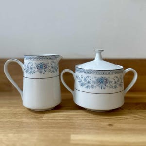 Może przedstawiać: Biały porcelanowy zestaw do śmietanki i cukiernicy z pokrywką. Oba elementy mają niebieski kwiatowy wzór ze srebrnym wykończeniem. Śmietanka ma zakrzywiony uchwyt, a cukiernica dwa uchwyty i pokrywkę z małym guzikiem.