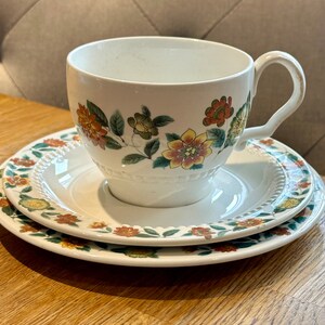 May include: A white teacup and saucer set with a floral pattern in shades of orange, yellow, green, and brown. The cup has a handle and the saucer has a scalloped edge.