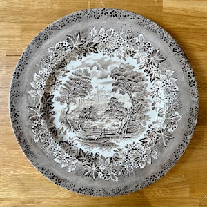 May include: A round ceramic plate featuring a brown and white transferware pattern. The center depicts a landscape with a castle, trees, and a body of water. The wide rim is decorated with a detailed border of leaves, berries, and small flowers.
