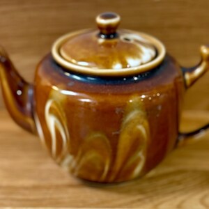 May include: A brown ceramic teapot with a lid, spout, and handle. The teapot has white and cream-coloured marbling. The lid has a small knob. The teapot is on a wooden surface.