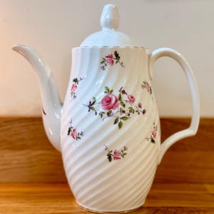 May include: White ceramic coffee pot with a lid and handle. The pot features a spiral design and is adorned with pink rose floral patterns and green leaves. The rim has a gold trim. The spout is curved and the handle is arched.