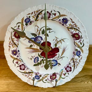 Puede incluir: Un reloj de pared decorativo hecho de un plato de cerámica blanco con un diseño floral. El plato presenta flores rojas, azules y moradas con manecillas doradas. El reloj está montado en un soporte metálico.