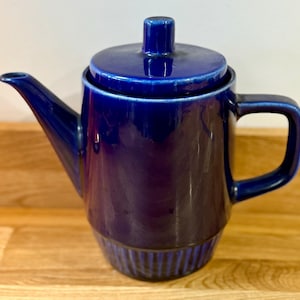 Könnte beinhalten: Eine dunkelblaue Keramik-Teekanne mit Deckel und Henkel. Die Teekanne hat eine glänzende Oberfläche und einen gerippten Boden. Der Ausguss ist lang und gebogen. Der Griff ist ein abgerundetes Rechteck. Der Deckel hat einen kleinen Knopf.