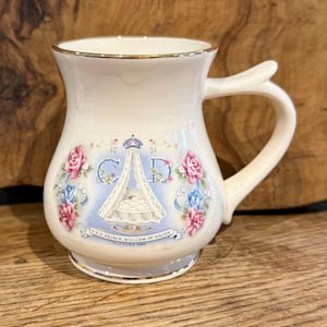 Puede incluir: Una taza de cerámica color crema con borde y base dorados. El frente presenta una ilustración central de una cuna bajo una corona, flanqueada por rosas rosas y azules. El texto dice H.R.H. PRINCE WILLIAM OF WALES 21ST JUNE 1982.