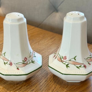 Peut inclure: Deux salières en céramique blanche avec un motif floral. Chaque salière présente un ruban rose et de petites fleurs roses avec des feuilles vertes. La base octogonale a une bordure verte. Le dessus a trois trous.