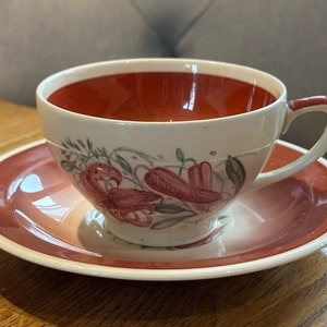 May include: A vintage teacup and saucer set with a floral design. The cup and saucer are white with a red rim and feature a pink floral pattern.