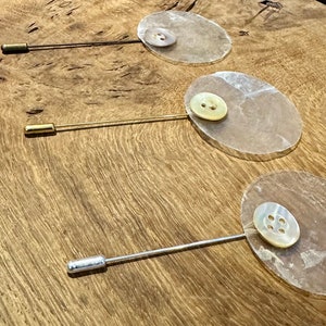 以下が含まれることがあります： 丸い半透明の白い貝殻のような土台が付いた装飾的なピンが3つあります。各土台の中央には異なるボタンが付いています。ピンは金、銀、ブロンズです。